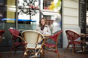 young smiling beautiful caucasian woman wearing a beige trench and brown scarf sitting at the table on the terrace of cafe.