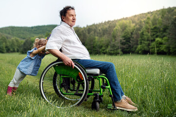 Small girl with senior grandfather in wheelchair on a walk on meadow in nature.