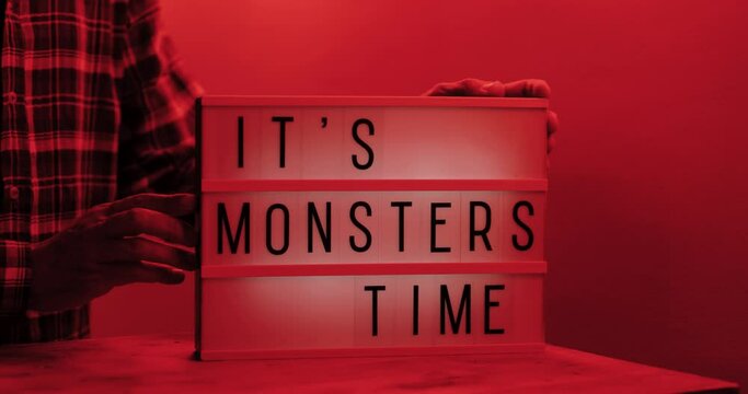 It's Monsters Time text displayed on a LED lightbox man tightening the box with both hands red light in the room, medium close-up, eye level, 4k, cinematic 
