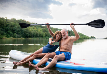 Senior couple sitting on paddleboard on lake in summer, taking selfie with smartphone.