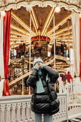 Young stylish girl in a fashionable clothes posing near a vintage carousel in the city. She relaxing in amusement park in winter.