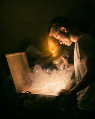 Young man in white t-shirt with a lamp in his hand, looking into a chest with smoke and mystical...