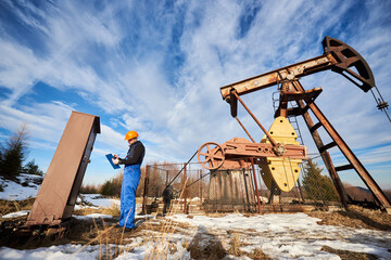 Petroleum engineer in work overalls and helmet making notes on clipboard while controlling work of...