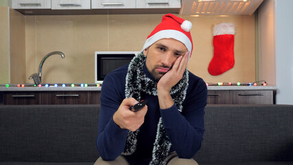 Tired bearded man in New Year red Santa Claus hat holds remote control in hands switches channels on TV. Male in blue sweater at home sits on couch on background Christmas kitchen watches television
