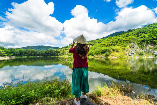 Vietnamese girl in dark red and bottle green traditional costume dress with conical hat standing in front of Ba Be lake
