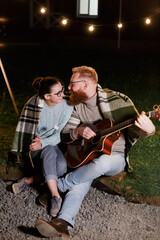 Young couple, caucasian woman and red haired bearded man, on a night picnic with a guitar and marshmello have a good time