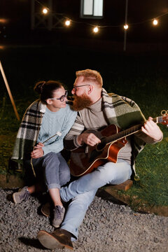 Young couple, caucasian woman and red haired bearded man, on a night picnic with a guitar and marshmello have a good time