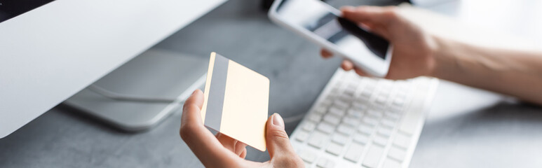 Panoramic shot of freelancer holding credit card and smartphone near computer