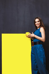 smiling brunette in denim jumpsuit holding a lemon in her hands near yellow rectangle she drew