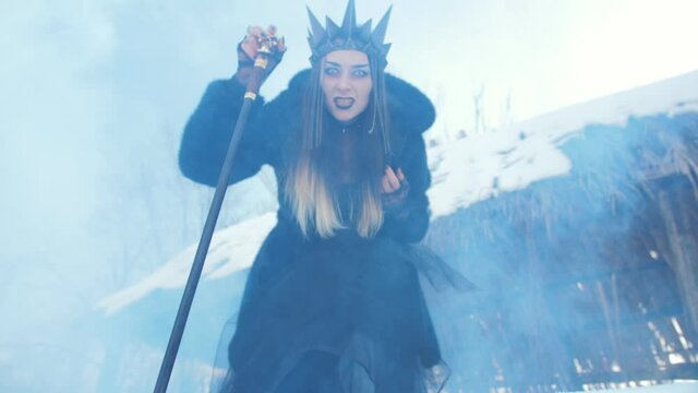 A scary witch near a wooden shack with crown on her head and a devilish cane in her hands is terrifying looking at the camera.