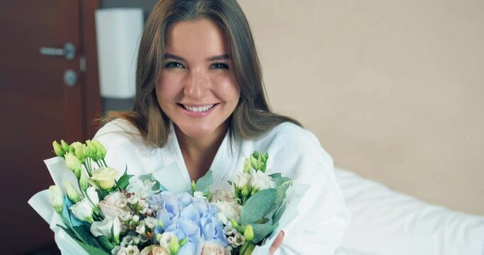 Smiling young lady in bathrobe looks at fresh flowers bouquet sitting on comfortable bed in hotel room in morning closeup