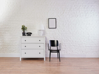 Decorative white cabinet and black chair in front of the brick wall, lamp, frame and vase of plant decorative style.