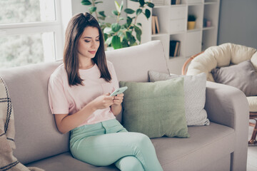 Photo portrait of woman holding phone in two hands sitting on sofa with crossed legs indoors