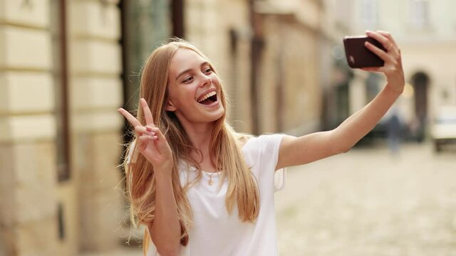 Young smiling hipster blond woman in white t-shirt posing, takes selfies and streams video to social media from her smartphone.