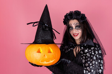 Halloween. A young woman in a costume and scary makeup with a carved Jack-o ' - Lantern pumpkin in a witch's hat. Isolate on a pink background