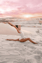 Fototapeta na wymiar young woman in white body is sitting in gymnastic pose in longitudinal twine on the sand in desert at sunset, fashion sport concept, free space