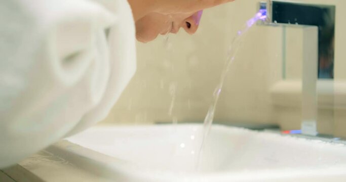 Young lady wearing white bathrobe washes hands and face with running water under tap with pink light in bathroom closeup