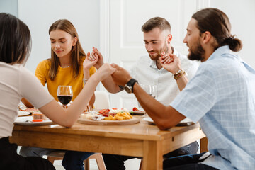 Image of happy multinational friends praying before lunch with holding hands together