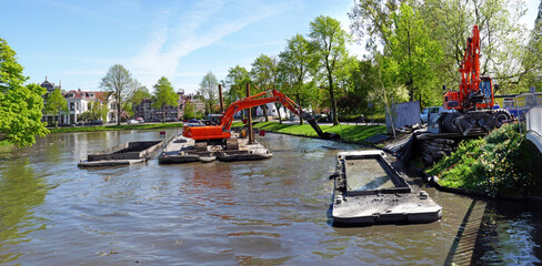 Dredging a city canal by crane from a pontoon; dredging spoil is transported in barges to a...
