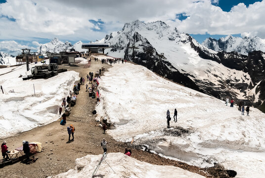 Rocks, Pink Snow, Mountain Sun Dazzles Eyes, Clouds, Blue Sky. Tourists Roam The Stony Path, Buy Souvenirs. Village Of Dambay 2750 M, Caucasus, Russia.