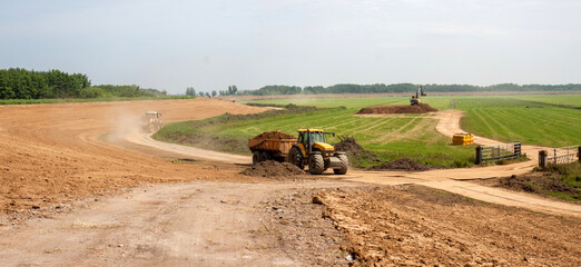 A tractor with tipper trailer deposits a load of clay on a large pile, neatly finished by a an excavator standing on top.