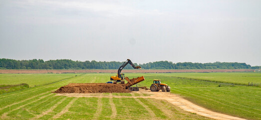 A tractor with tipper trailer deposits a load of clay on a large pile, neatly finished by a an excavator standing on top.