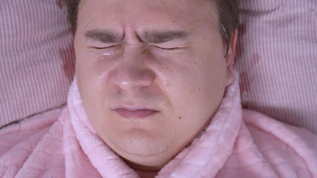 Portrait of sleeping man in funny pink pajamas who has terrible nightmare and so he is drenched in streams of cold sweat on face and mumbles in sleep, top view, close up.