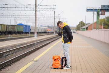 Young man stands on platform, waiting for train. Male passenger with backpacks on railroad platform in waiting for train ride. Concept of tourism, travel and recreation.