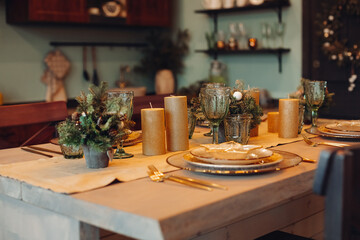 Close-up of beautifully decorated dining table with fir trees in vases and gold candles. Sets of plates for four people.