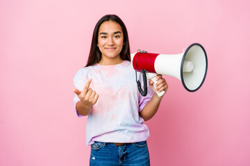 Young asian woman holding a megaphone isolated on pink background pointing with finger at you as if inviting come closer.