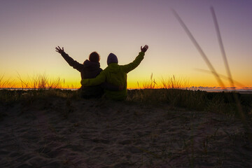 Junges Pärchen blickt glücklich mit erhobenen Händen auf Sonnenaufgang in den Dünen