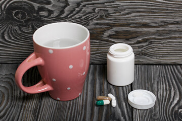 Container with pills and a mug of water. The lid of the container and several tablets are next to each other. On pine planks in black.
