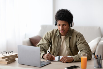 Studying from home. African American teenage guy in headphones writing down info during online...