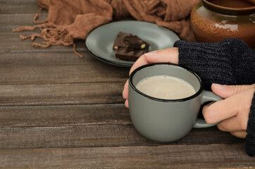 Saucer and dark chocolate on it, brown scarf on the wooden table and woman holding grey cup of milky chocolate drink.Empty space
