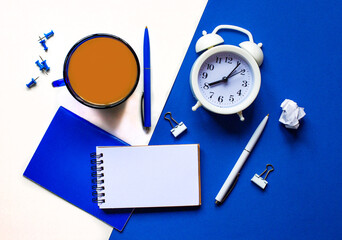 On a white and blue background, a white alarm clock, a pen, a notebook, a blue cup with coffee, a blue pen and buttons. Flat lay