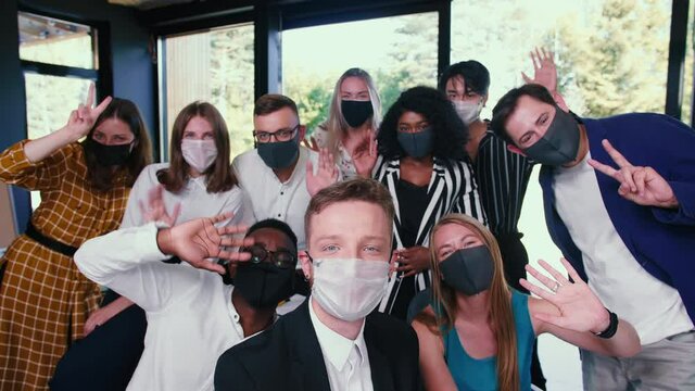 Group of happy diverse business colleagues posing, waving for selfie photo in masks against COVID-19 at modern office.
