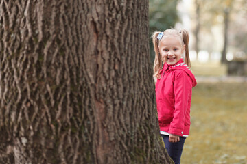 A little girl with two ponytails on her head stands near a wide tree trunk in the park