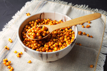 Sea buckthorn in a clay bowl with a wooden spoon on a dark table, close-up.