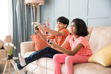 Cheerful Asian Indian kids playing video game using controller or joystick, sitting on sofa, couch © StockImageFactory