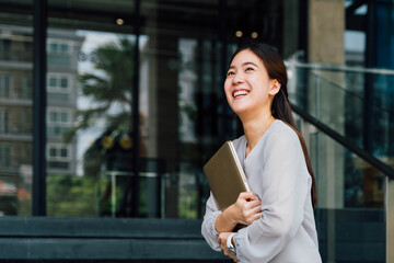 Fototapeta premium One successful and happy young adult business woman holding a laptop with smile. Professional female worker standing in corporate modern office and window in casual gray look. - With copy space.