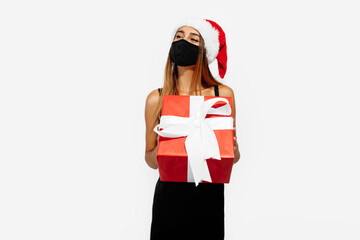 Young woman wearing red christmas hat and medical protective mask on her face holding gift box on white background, concept of christmas, coronavirus