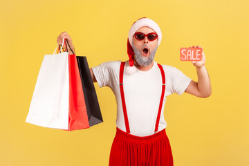 Excited surprised man in santa claus hat showing and holding paper shopping bags and sale card, looking at camera with shock, holidays discount. Indoor studio shot isolated on yellow background