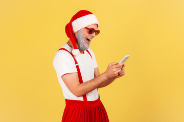 Happy funny man in santa claus costume and stylish sunglasses typing on smartphone and smiling, reading pleasant news. Indoor studio shot isolated on yellow background