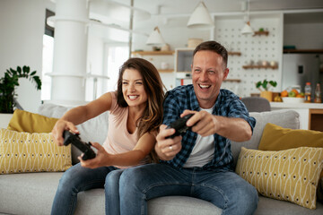 Husband and wife playing video game with joysticks in living room. Loving couple are playing video games at home. © JustLife