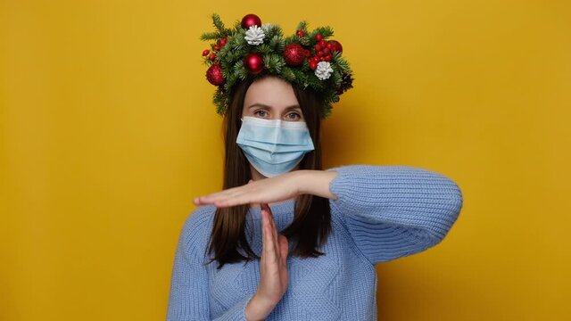 Dissatisfied young woman in medical face mask, shows timeout gesture, needs stop, wears handmade wreath and blue sweater, isolated on yellow studio wall. Pandemic coronavirus and Christmas concept