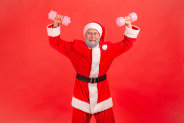 Fototapeta na wymiar Positive funny elderly man in santa claus costume and eyeglasses raising dumbbells and fooling face, preparing for winter holidays. Indoor studio shot isolated on red background