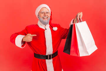 Smiling elderly gray bearded man in santa claus costume pointing finger at paper shopping bags in his hand, doing holidays purchase. Indoor studio shot isolated on red background