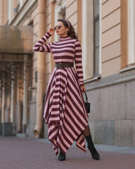 Elegant woman in long warm dress with pink and brown stripes walking at city street. Female model...