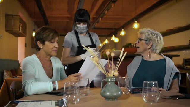 Two happy senior women with the menu in hands ordering food at a restaurant. Young waitress with protective face mask and gloves taking an order. Food and drinks during Coronavirus outbreak.