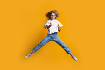 Fototapeta na wymiar Jumping blonde woman is gesturing the shot and gun with both hands while jumping on a yellow wall at studio